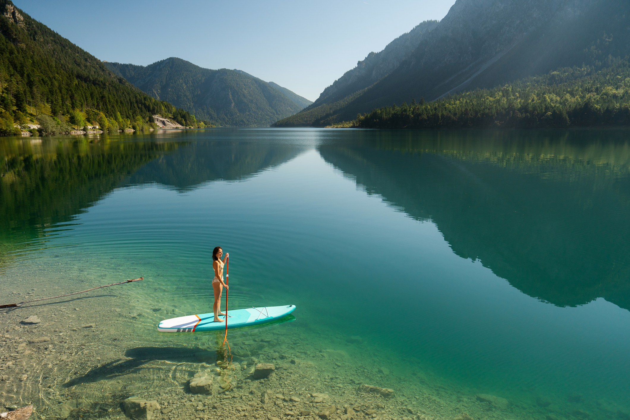 Préparer son séjour | GettyImages 2155528448 Woman SUP boarding on mountain lake in Alps surrounded by pine forest